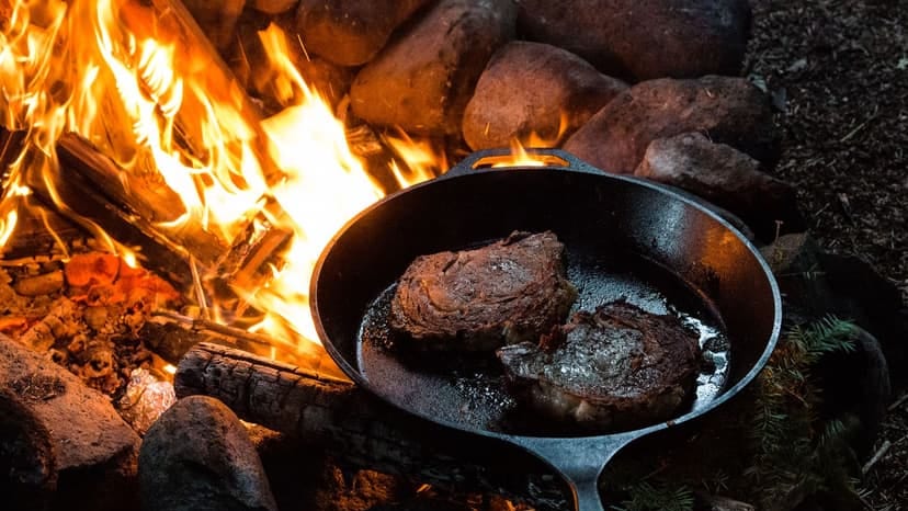 Sizzling steaks cooking in a cast iron pan over a campfire surrounded by logs and rocks.