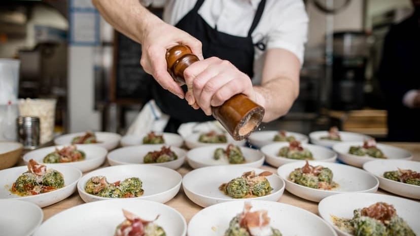 Bearded chef seasoning gourmet dishes with pepper in a busy restaurant kitchen.