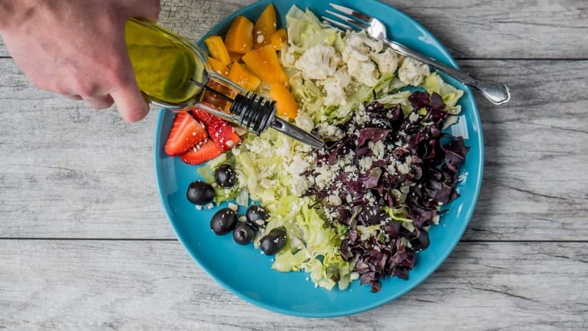 A vibrant vegetable salad with olives, feta, and strawberries being drizzled with olive oil.
