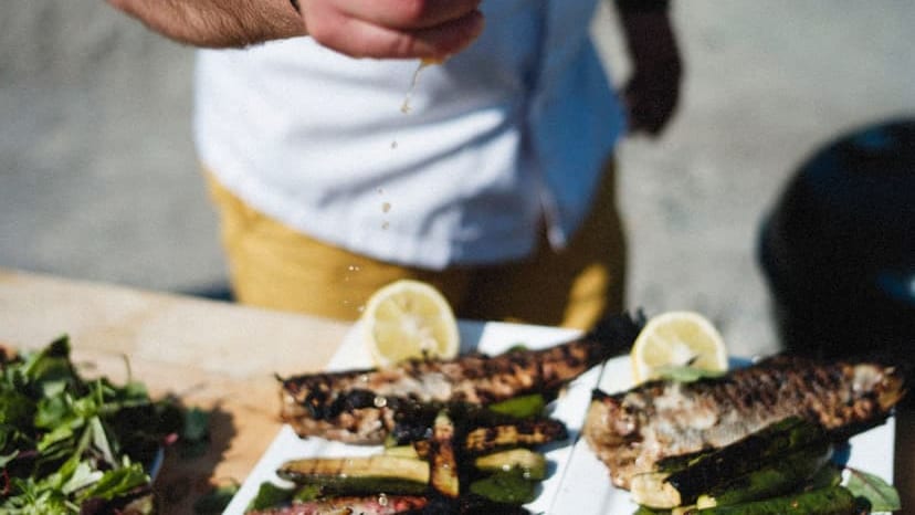 A person is squeezing lemon over grilled fish and vegetables on a sunny day.